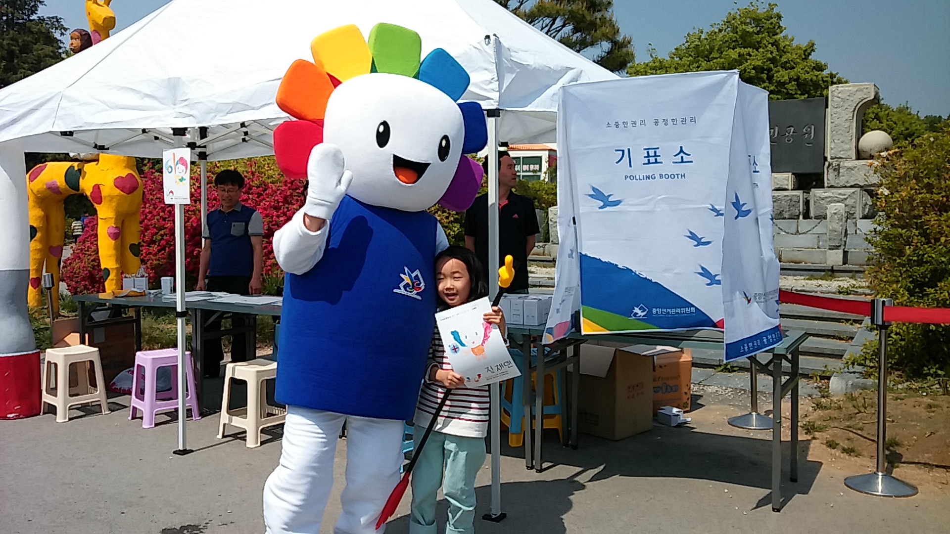 선거관리위원회 캐릭터 참참이 인형과 그림을 든 어린이가 함께 서 있고 뒤편으로는 홍보 부스가 차려져 있으며 오른편에는 기표소가 있는 사진