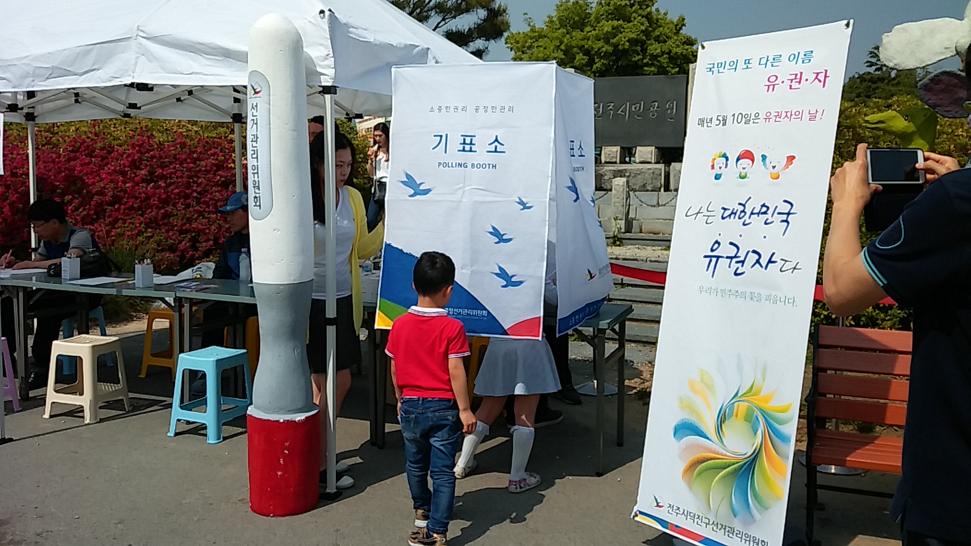 대형 기표용구 모양의 조형물이 왼쪽에 있고, 오른쪽에는 유권자의 날 배너가 있으며, 배너에는 '국민의 또다른 이름 유권자, 매년 5월 10일은 유권자의 날! 나는 대한민국 유권자다. 우리가 민주주의의 꽃을 피웁니다'라고 적혀있음. 화면 가운데에는 기표소가 있는데 기표소 안에서 한 어린이가 모의투표 체험을 하고 있으며, 다른 아이는 차례를 기다리며 줄을 서 있는 모습