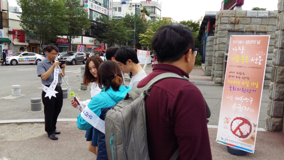 '5월 18일 성년의 날, 가장 기분좋은 선물, 성인으로서 유권자가 되신 것을 축하드립니다. 내가 처음으로 하게 되는 선거는 2016년 4월 13일 국회의원선거, 나는 대한민국 유권자다.' 라고 적힌 배너가 놓여 있고, 이를 바라보고 있는 남학생의 모습