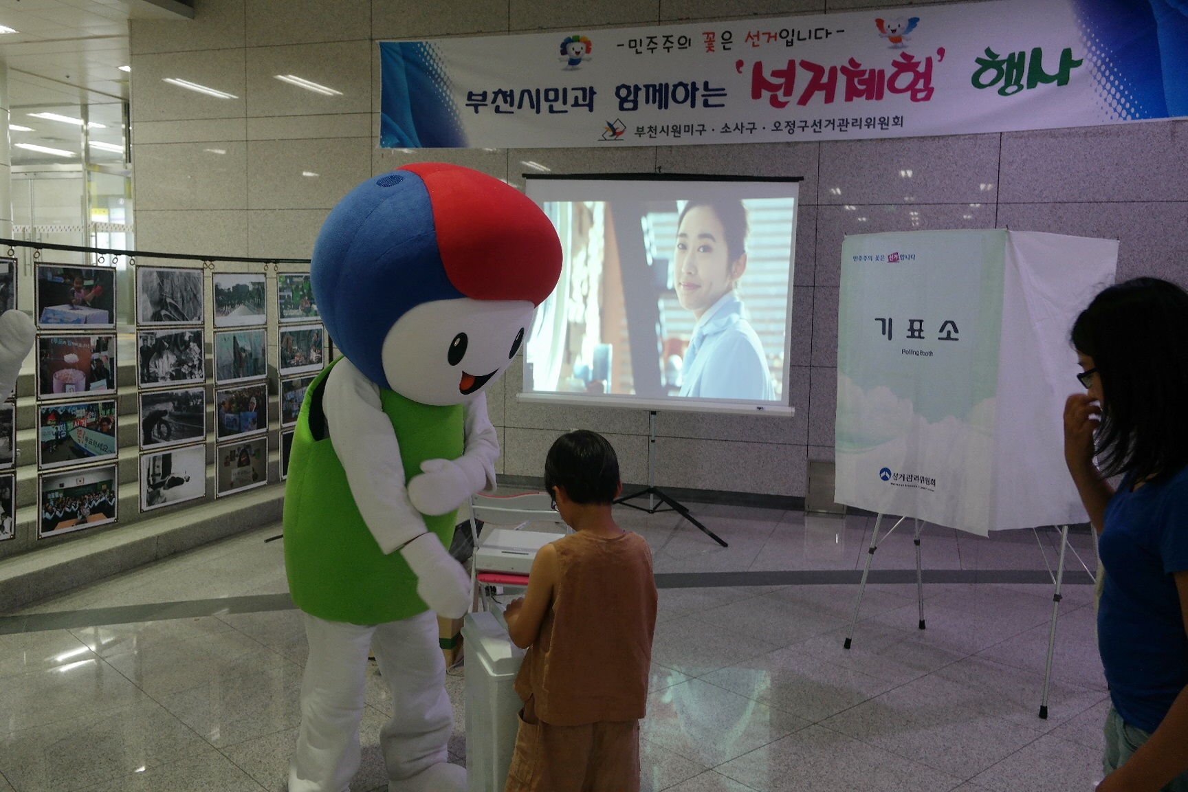 사진전시대와 사전투표홍보영상을 배경으로 "바루"의 안내에 따라 어린이가 투표함에 투표용지를 투입하고 있는 모습입니다.