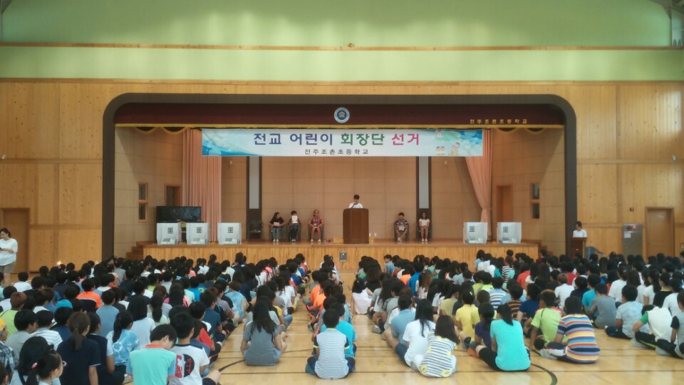 강당 단상 위쪽에 전교 어린이 회장단 선거 라는 현수막이 붙어 있고 단상 위에는 후보자 학생들이 앉아 있으며 한 후보자가 정견을 발표하고 있는 모습