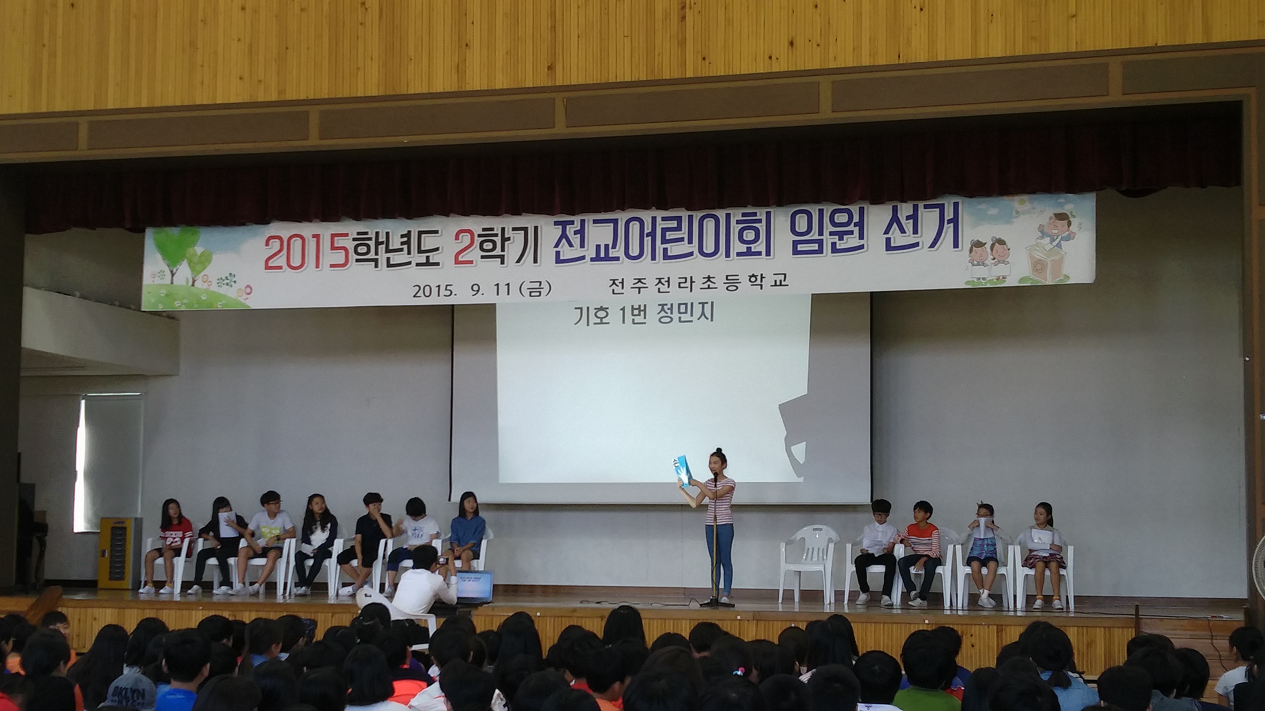 2015학년도 2학기 전교어린이회 임원 선거라는 현수막이 단상 위에 붙어 있고, 단상에는 후보 학생들이 앉아 있으며, 이 중 한 후보자가 정견발표를 하고 있는 모습
