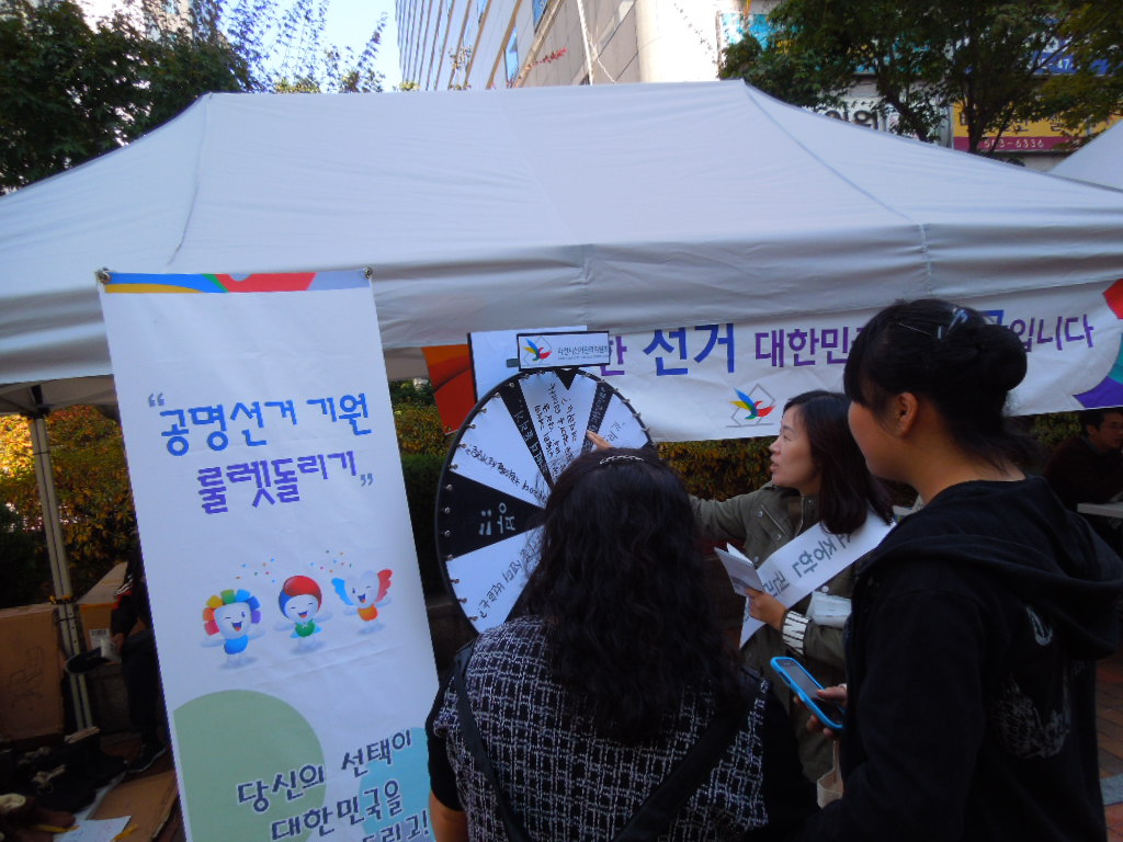 과천시 주말장터 내 행사부스에 설치된 공명선거기원 룰렛을 돌리고 퀴즈를 풀고 있는 시민의 모습
